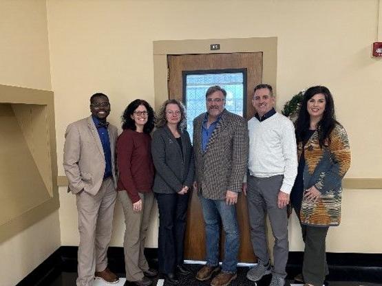 a group of six people smiling at the camera - in the background is a door and beige wall